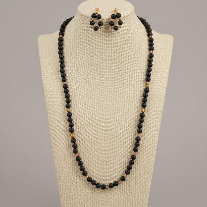 A black beaded necklace and matching stud earrings displayed on a black jewelry bust. The beads are primarily black with small gold-toned accents, set against a patterned tablecloth and a red-orange wall in the background.