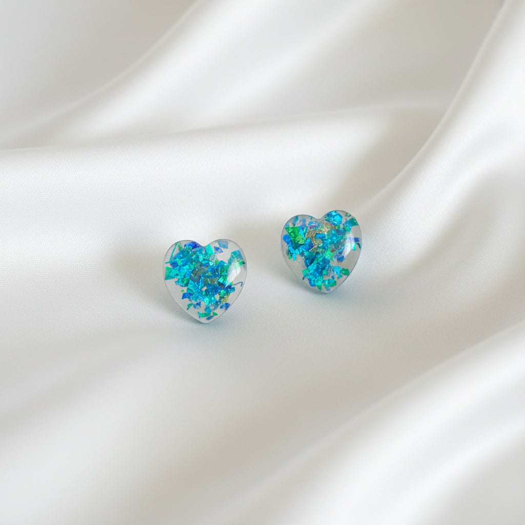 A pair of heart-shaped stud earrings with a vibrant blue, green, and orange iridescent opal pattern, displayed on a black velvet card inside a black plastic sleeve, set against a dark brown wooden background.