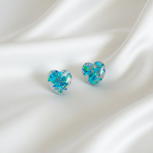 A pair of heart-shaped stud earrings with a vibrant blue, green, and orange iridescent opal pattern, displayed on a black velvet card inside a black plastic sleeve, set against a dark brown wooden background.