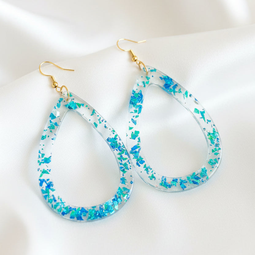 A pair of teardrop-shaped dangling earrings with iridescent blue and green glitter inside a clear resin-like border, hanging from gold-tone fishhook ear wires on a dark brown wooden surface.