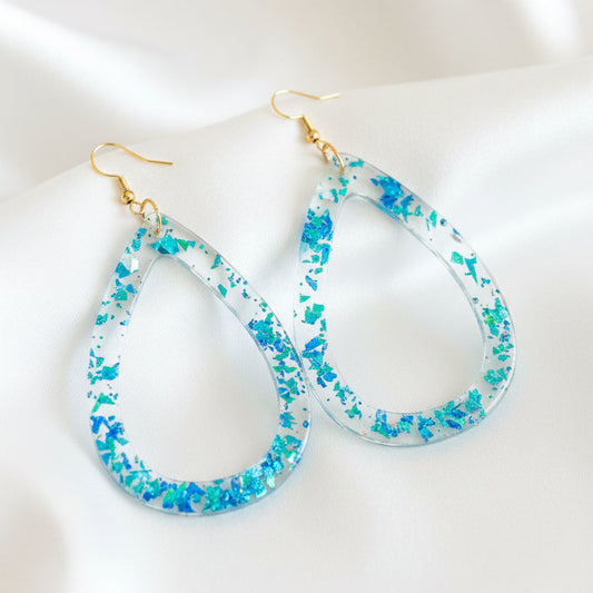A pair of teardrop-shaped dangling earrings with iridescent blue and green glitter inside a clear resin-like border, hanging from gold-tone fishhook ear wires on a dark brown wooden surface.